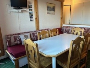 Un comedor con una mesa rectangular y sillas de madera, un televisor montado en la pared y un cuadro enmarcado sobre la mesa.