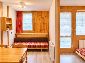 A cozy room with a wooden floor, a bed with a plaid bedspread, a wooden table, a shelf with a plant, and a window with a red curtain.
