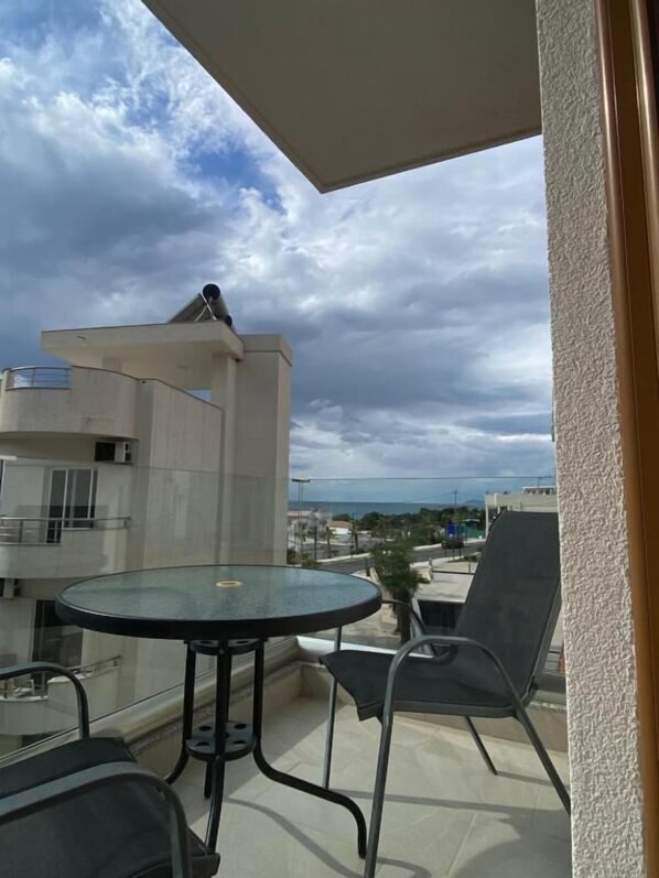 Un balcón con una mesa y sillas de vidrio, con vistas a una zona residencial y al mar bajo un cielo nublado.