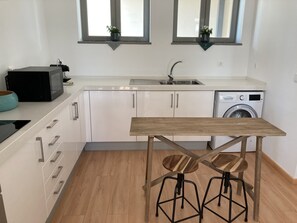 Uma cozinha moderna com armários brancos, uma máquina de lavar Bosch, uma mesa de madeira com banquetas e duas janelas com plantas.