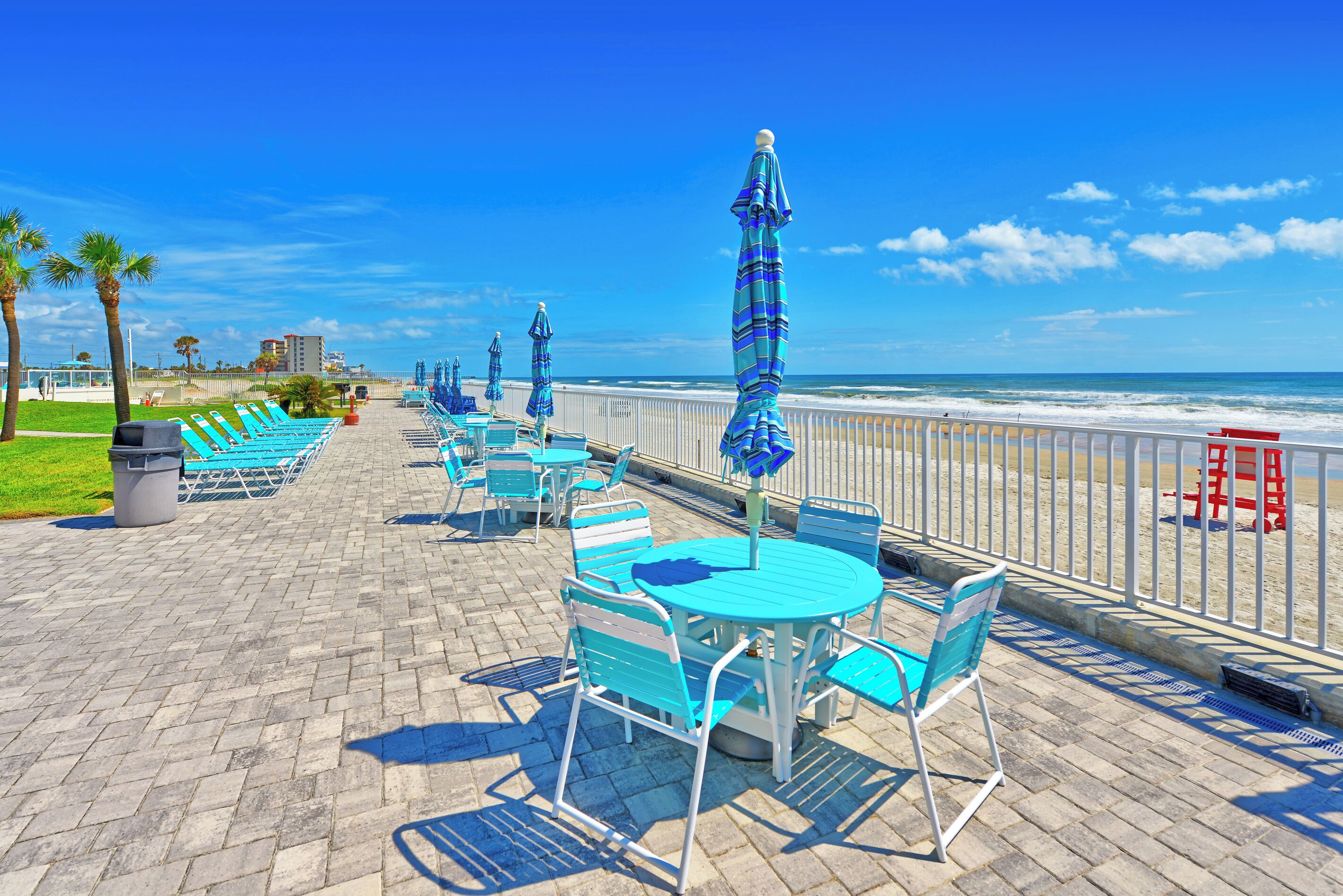 Une promenade en bord de mer avec des tables et des chaises à l’extérieur, des parasols, une plage en arrière-plan et un ciel dégagé.
