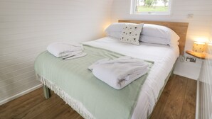 Un lit bien fait, avec du linge de lit blanc et un plaid vert, une lampe de chevet et une fenêtre donnant sur des arbres.
