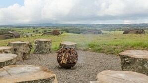 Un espace extérieur rustique aménagé avec des sièges, doté d’un foyer sphérique original, entouré d’escaliers en pierre et offrant une vue panoramique sur des collines ondoyantes et des bâtiments éloignés.