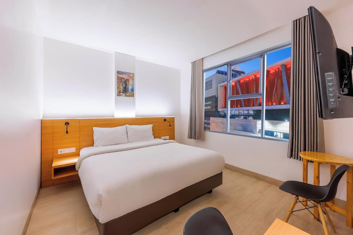 A hotel room with a bed, a desk, a chair, a TV, and a window with a view of a building.