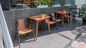 An outdoor seating area with wooden tables and wicker chairs, a glass wall reflecting buildings and greenery, and a tiled floor. - GRAND   THAMRIN  HOTEL (Jakarta)