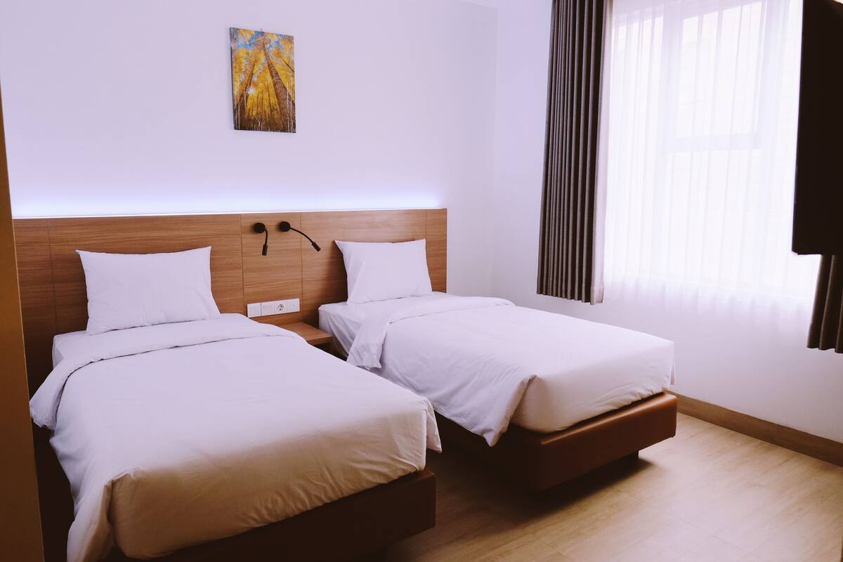 A hotel room with two single beds, a wooden headboard, a bedside table, a nightstand, a wall-mounted lamp, and a window with curtains.