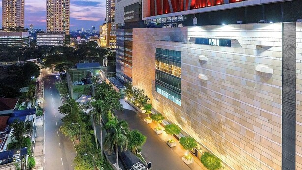 A modern building with a unique red structure on the roof, surrounded by a well-maintained urban street with palm trees and a clear sky. - GRAND   THAMRIN  HOTEL (Jakarta)