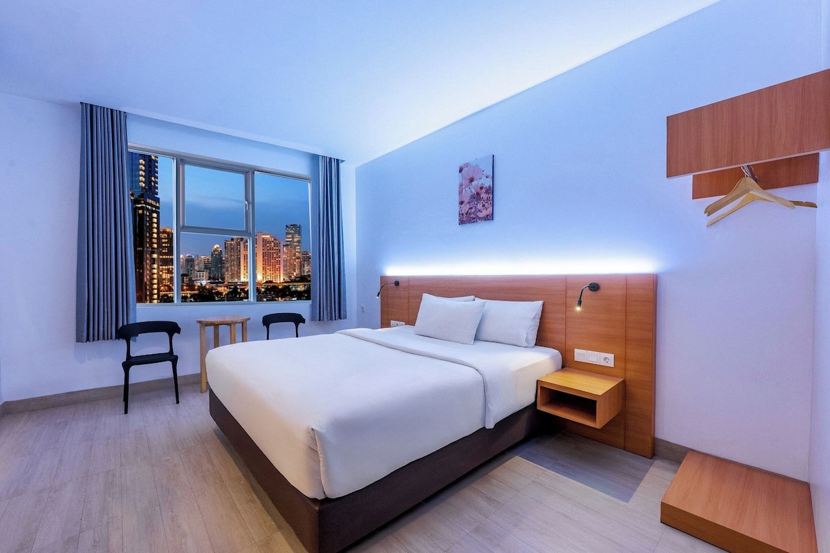 A modern hotel room with a large bed, a nightstand, a small table, a chair, and a view of a cityscape at dusk.