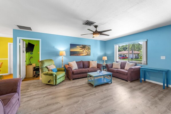 A living room with a blue wall, two couches, a coffee table, a ceiling fan, and a window with a view of houses. - Creative Coastal Cottage - 5 min to beach (Venice)