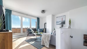 A modern living room with a sofa, a coffee table, and a fireplace. There is a large window with curtains, a painting of a lighthouse, and a small plant on the wall. - Sloop-Lounge - Fireplace, Infrared Sauna, Bathtub, Shower, Balcony, 2 Floors (Olpenitz)