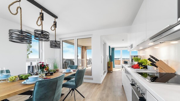A modern kitchen with a built-in stove, a dining area with a wooden table and blue chairs, and a view of the outdoors through large windows. - Sloop-Lounge - Fireplace, Infrared Sauna, Bathtub, Shower, Balcony, 2 Floors (Olpenitz)