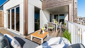 A modern outdoor patio with a wooden deck, a seating area with a table and chairs, and a sliding glass door leading to an interior space. - Sloop-Lounge - Fireplace, Infrared Sauna, Bathtub, Shower, Balcony, 2 Floors (Olpenitz)