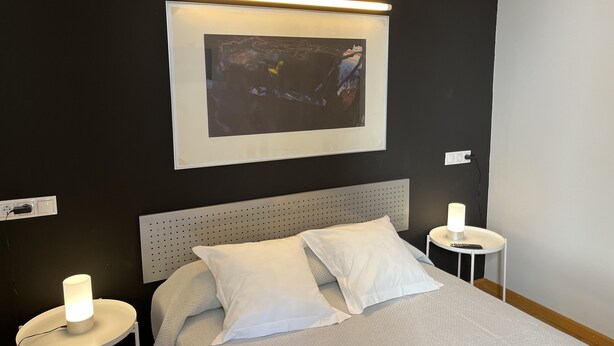 A neatly made bed with white bedding and pillows, a headboard, and a framed artwork above it. There are two bedside tables with lamps. - León Apartments. 20 years of success. Centrally located and recently air-conditioned (León)