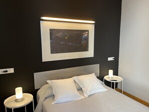 Un lit bien fait, avec une literie et des oreillers blancs, une tête de lit et une œuvre d’art encadrée au-dessus. Il y a deux tables de chevet avec des lampes.