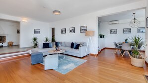 Amplia sala de estar con un sofá de gris claro, una mesita de centro y un comedor con mesa y sillas. Se observan varias fotografías enmarcadas en las paredes, y una lámpara es visible.