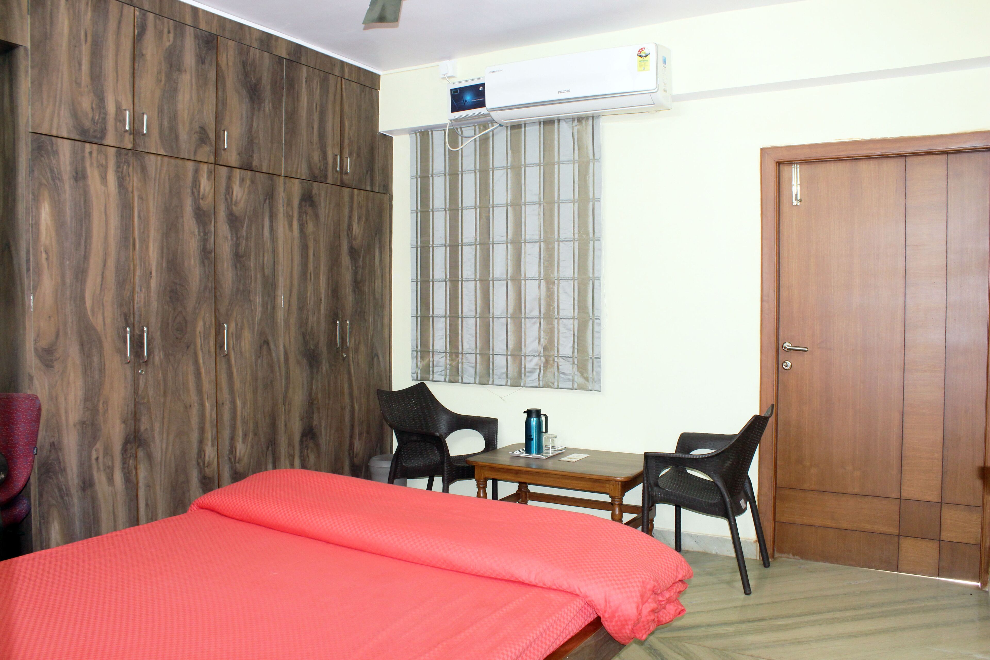 A bedroom with a bed, wardrobe, two chairs, a table with a kettle, and a window with blinds.