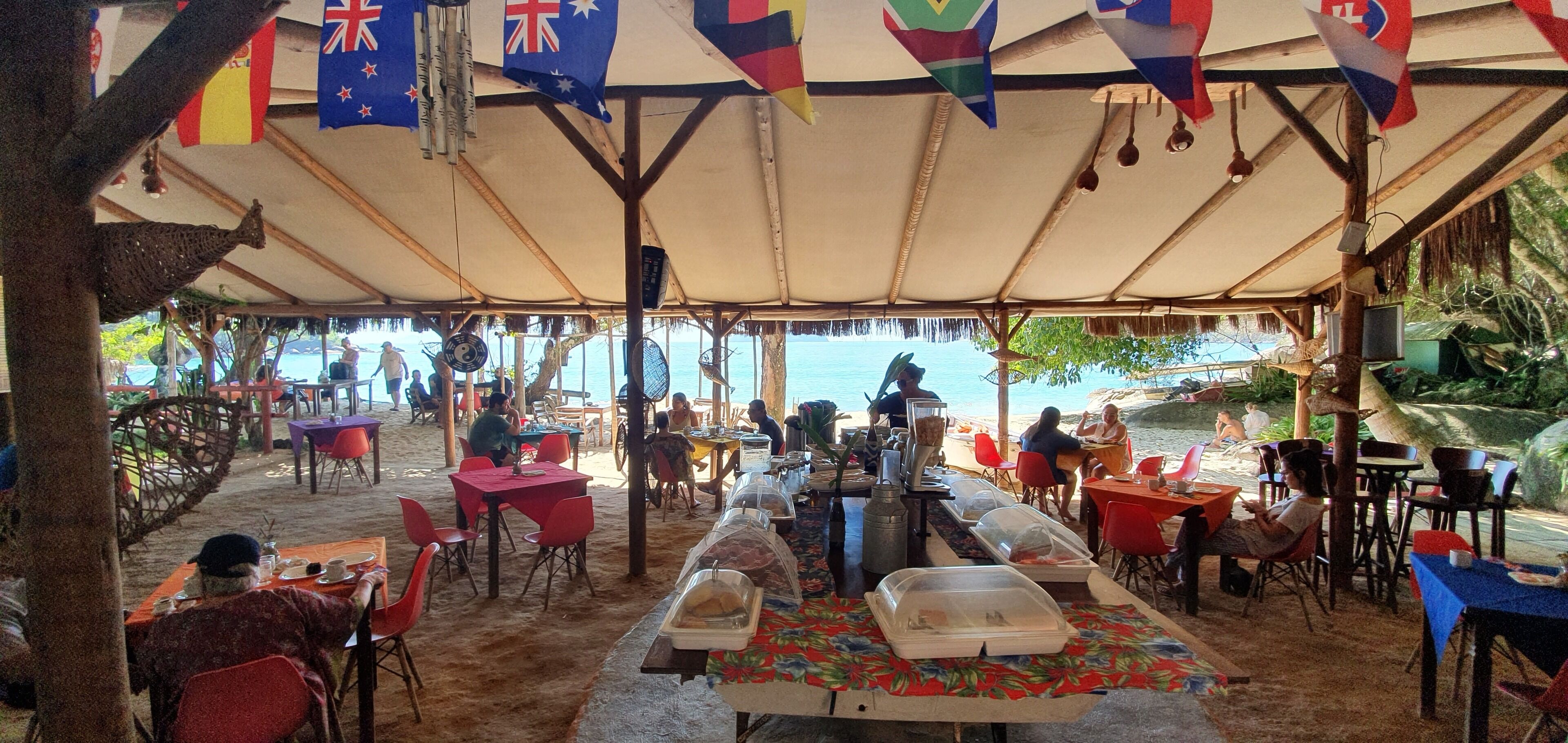 An outdoor dining area with tables and chairs, flags hanging above, and a view of the sea.