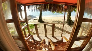 A beach view from a wooden deck with a thatched roof, featuring a sandy beach, palm trees, and the ocean. - Cabanas Paraiso (Angra dos Reis)
