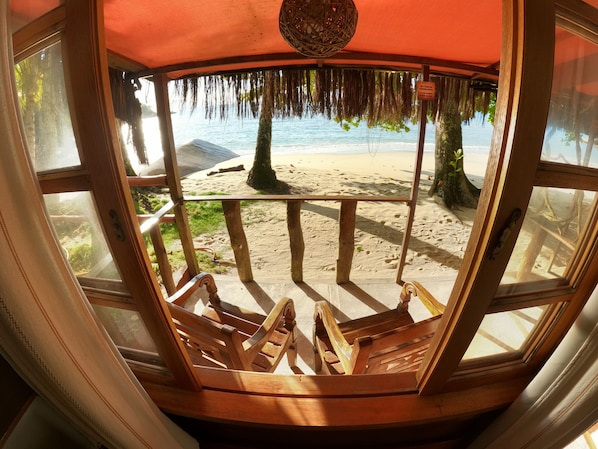 A beach view from a wooden deck with a thatched roof, featuring a sandy beach, palm trees, and the ocean. - Cabanas Paraiso (Angra dos Reis)