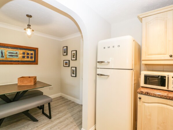 A compact kitchen with a white refrigerator, a microwave, and wooden cabinets. There is a dining table with a bench and a framed artwork on the wall. - The Beach House (Bridport)