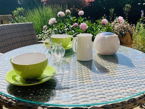 Une table ronde en verre, dont le bord est orné d’un motif tressé, est placée à l’extérieur, accompagnée d’une chaise en osier.