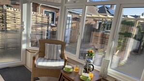 Un patio iluminado por el sol con mobiliario de mimbre, una mesa con una tetera, tazas y un vaso de jugo de naranja.
