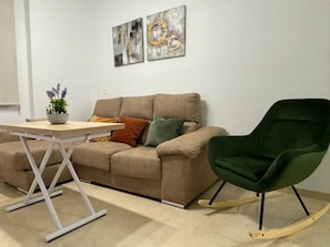 A room with a beige sofa, a green rocking chair, a small table, and two abstract paintings on the wall.