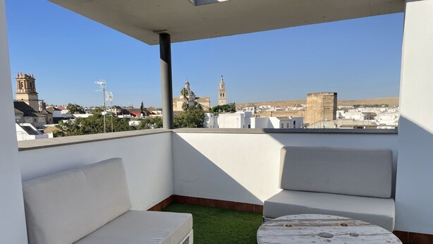 Eine Dachterrasse mit weißer Couch, einem kleinen runden Tisch und Blick auf die Stadtlandschaft, darunter ein Kirchturm.