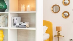 Ein modernes Zimmer mit weiß-gelber Farbgestaltung, das ein Bücherregal mit Büchern und Dekorationen, ein gelbes Bett und einen kleinen runden Tisch mit einer Lampe zeigt.