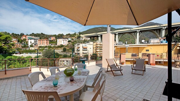 Eine Terrasse mit Tisch, Stühlen und Sonnenschirm, mit Blick auf eine Stadtlandschaft mit Gebäuden und Hügeln.