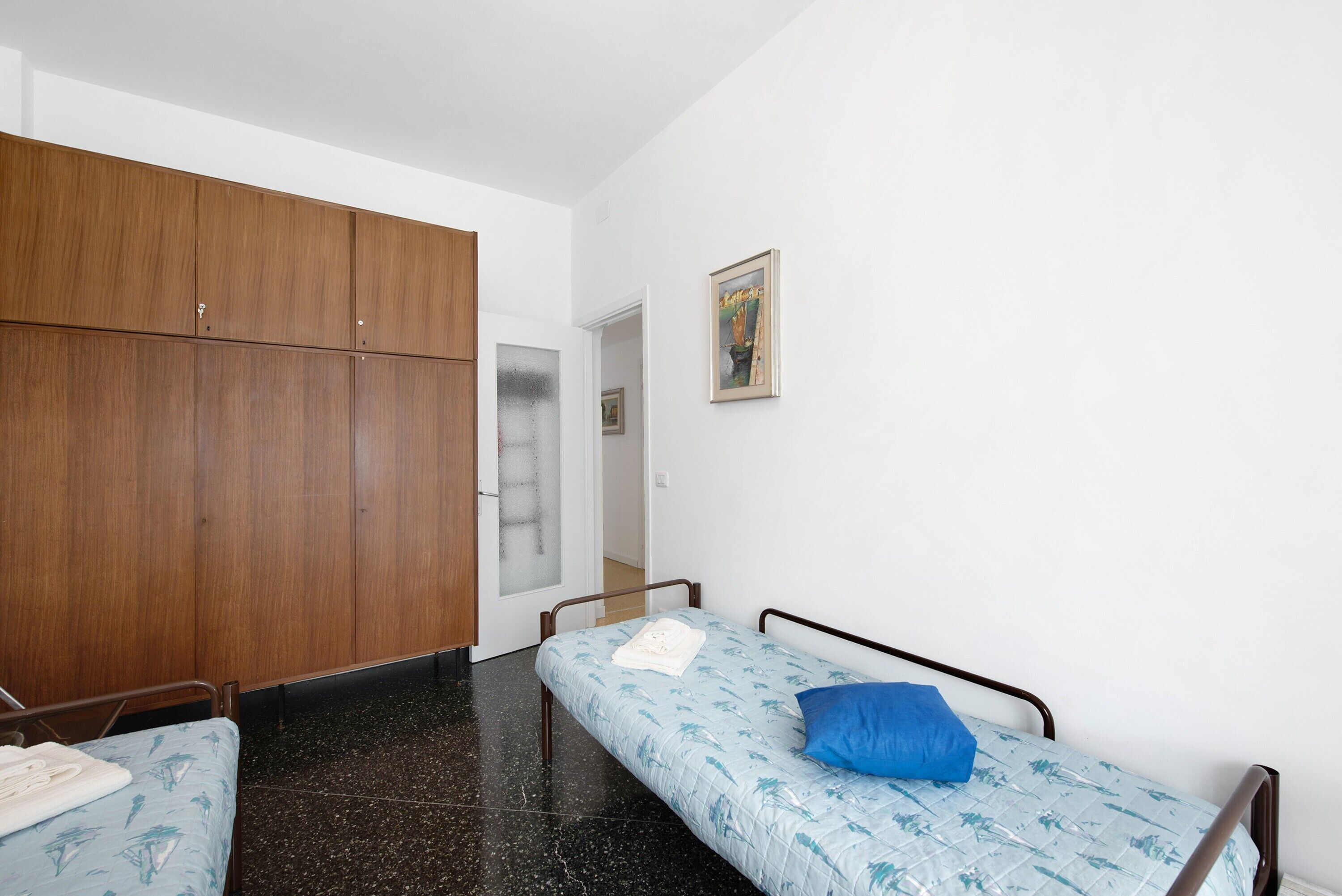 Ein Zimmer mit zwei Einzelbetten, einem Holzschrank, einer Tür zu einem anderen Raum und einem gerahmten Bild an der Wand.