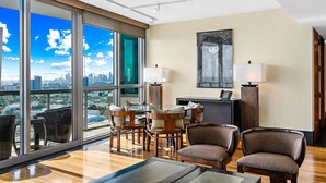 Quarto de hotel moderno com uma janela ampla que oferece vista para a cidade, área de refeições com mobiliário de madeira e uma TV de tela plana sobre um suporte.