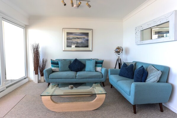 A living room with a blue sofa, a glass coffee table, and a framed picture on the wall. - Tides - Beach Front Apartment in Bracklesham Bay (Chichester)
