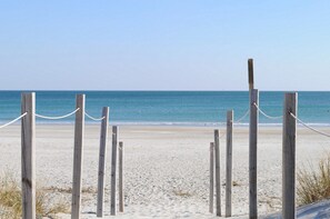 Ein Holzzaun mit einer Seilsperre führt zu einem Sandstrand, im Hintergrund das Meer.