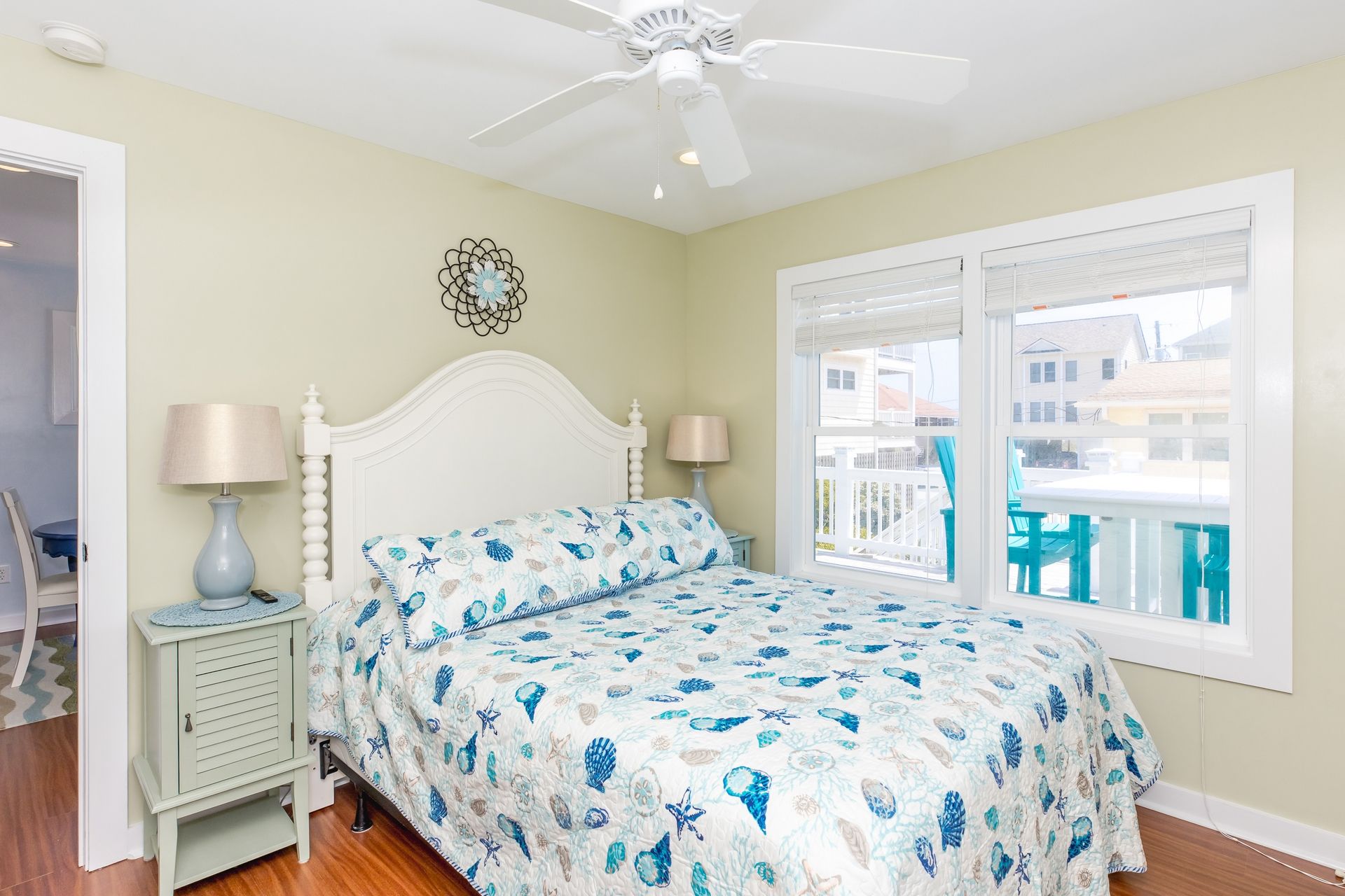 A bedroom with a bed, two bedside tables, a ceiling fan, a window with a view, and a decorative wall piece.