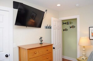 A room with a flat-screen TV mounted on the wall, a wooden dresser with drawers, a door with fish-shaped decorations, and a lamp on a small table. - Playin Hooky - Charming 3 Br/2 Ba Cottage!  Linens included for weekly stays! (Topsail Beach)