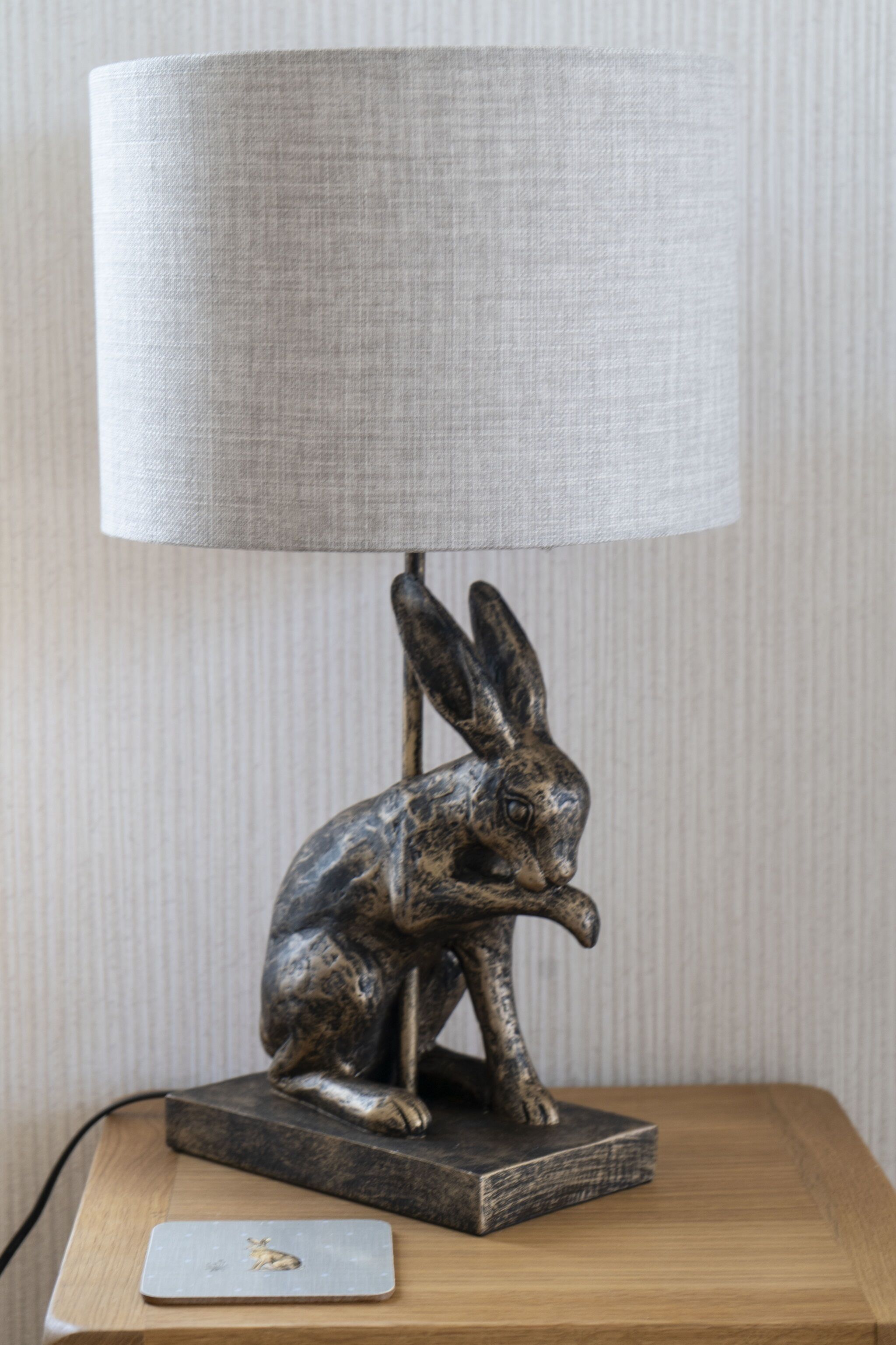 Una lámpara con base de escultura de conejo metálico y pantalla de tela sobre una mesa de madera.
