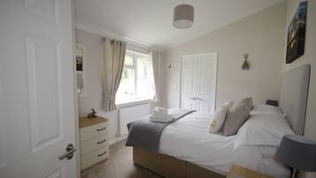 A bedroom with a bed, a bedside table, a lamp, a window with curtains, a radiator, and a door.