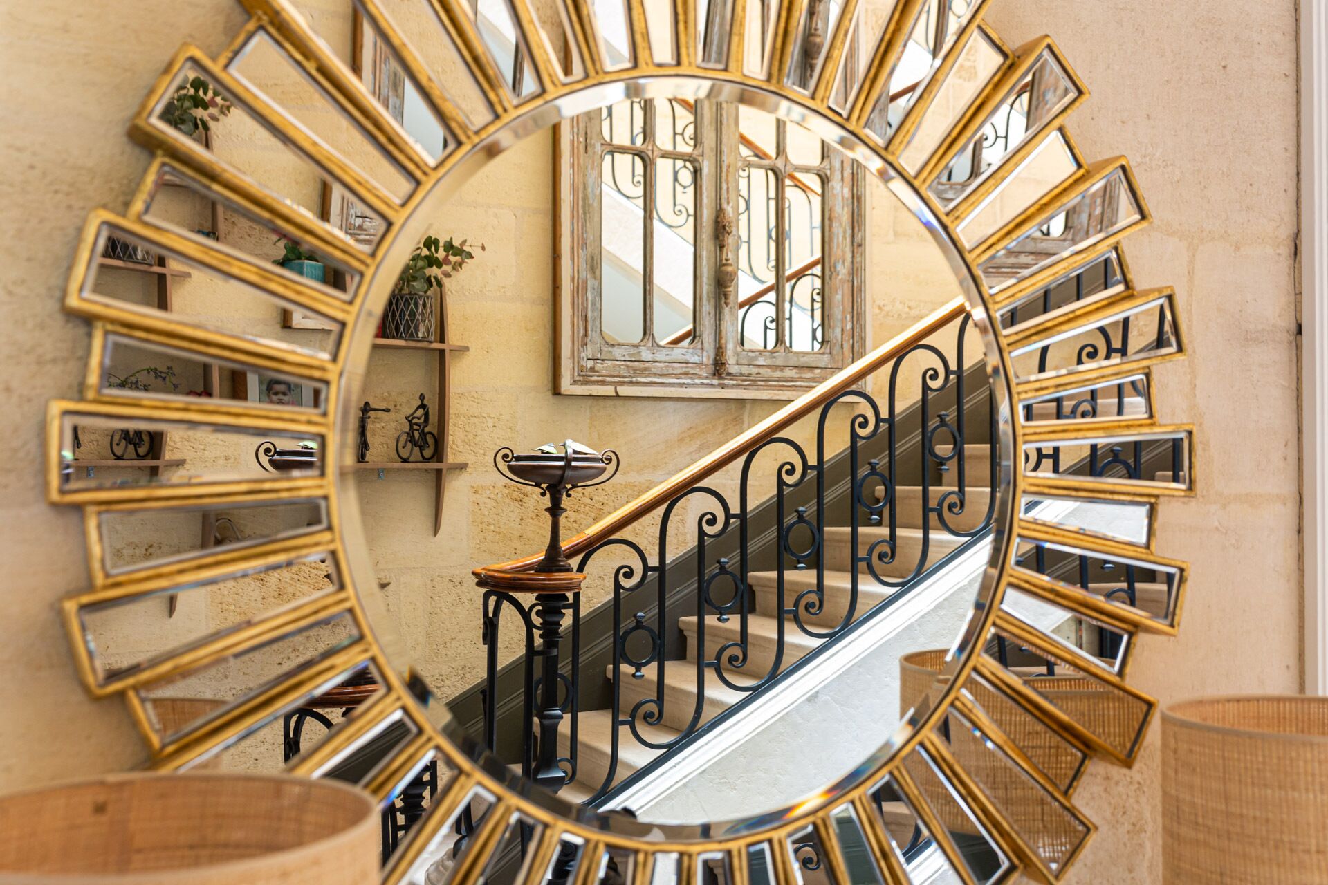 A sunburst mirror reflecting a staircase with a wrought iron railing and a window in the background.