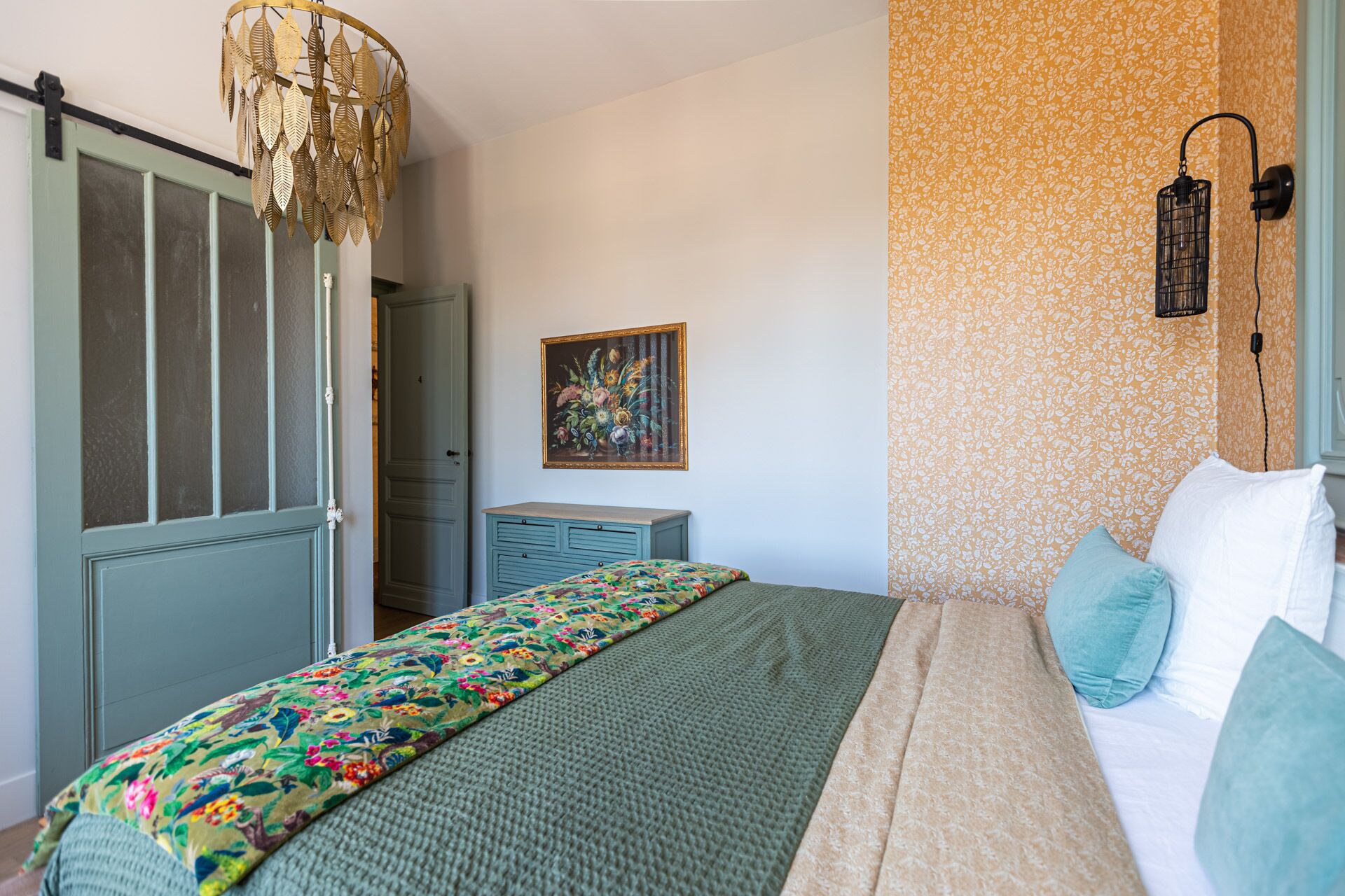 Een slaapkamer met een bed, een linnenkast, een deur, een wandlamp en behang met een bloemmotief.