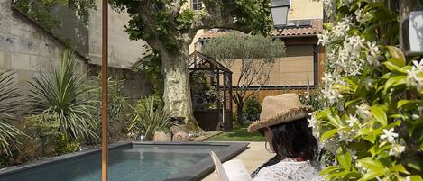 A poolside area with a chair, a thatched umbrella, and a person reading a book.
