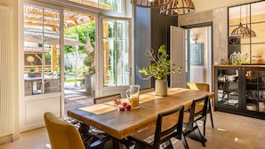 A dining area with a wooden table, chairs, and a vase with greenery. There are windows with a view of the outdoors. - La Maison Mulato Bordeaux, Saint-emilion (Libourne)