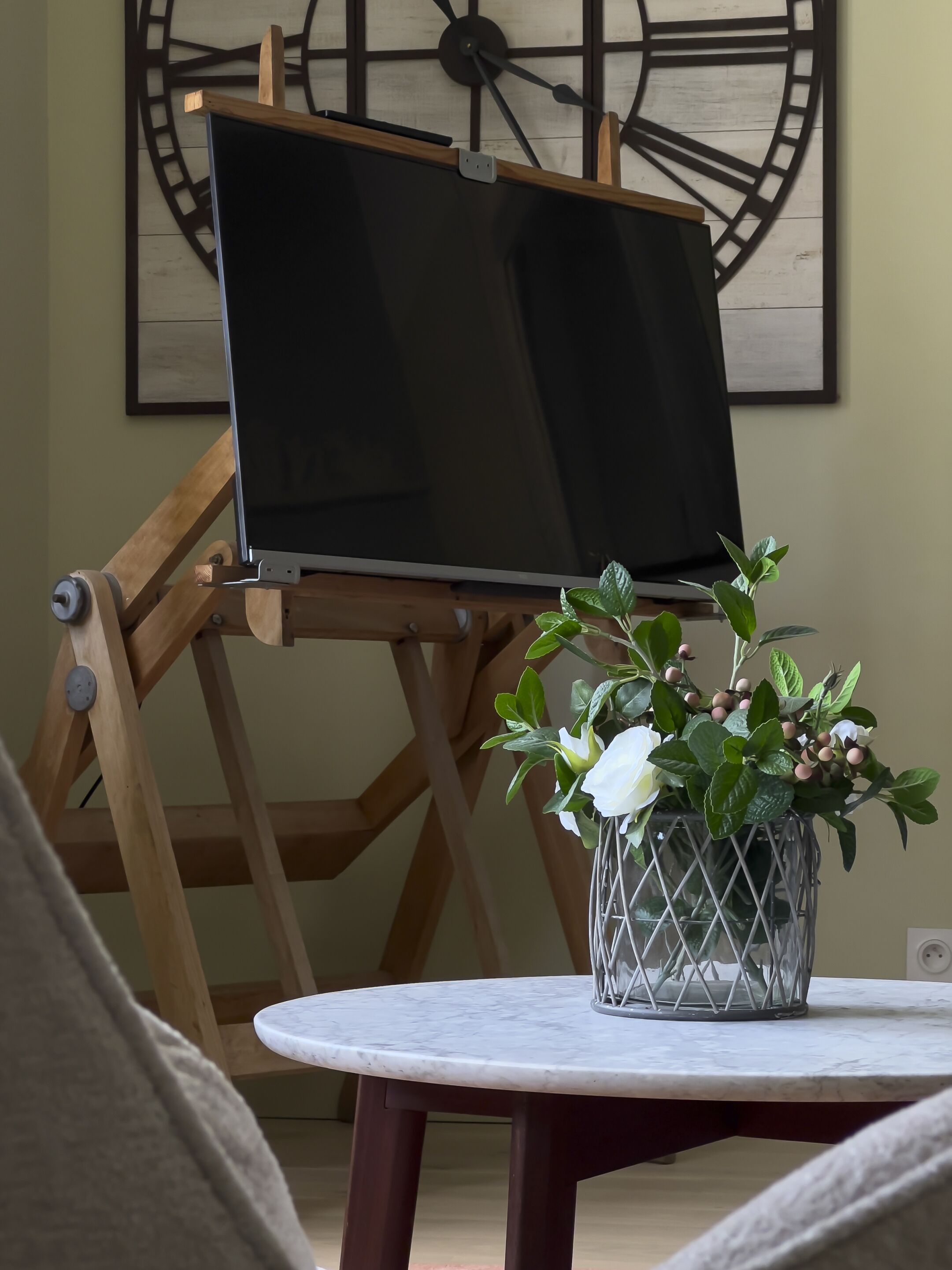 A room with a wooden easel holding a blank canvas, a potted plant with white flowers, and a large clock on the wall.