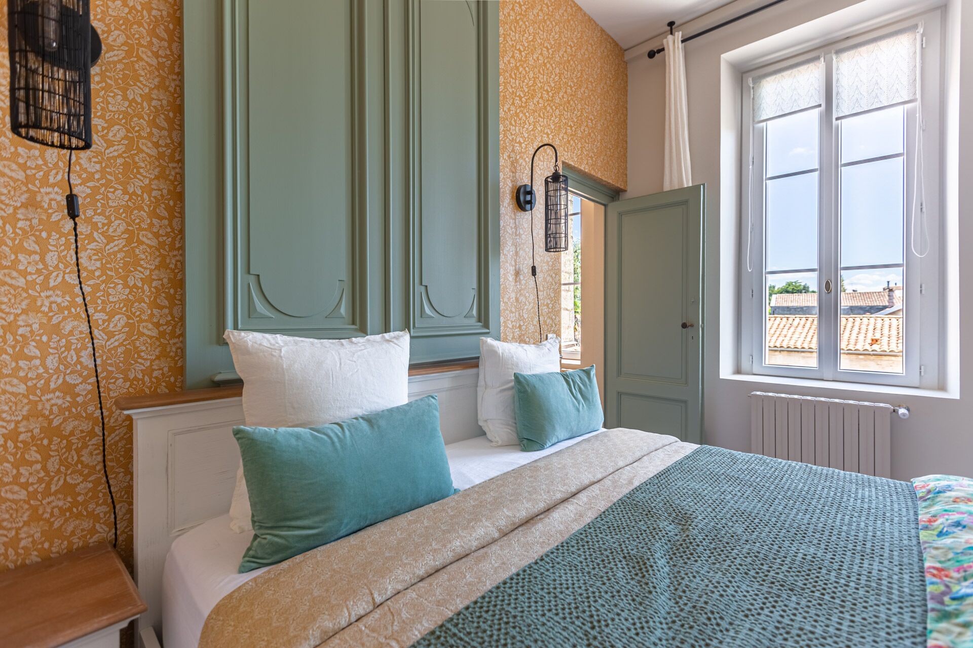 A bedroom with a bed, pillows, a window with a view, and a wall with a floral pattern.