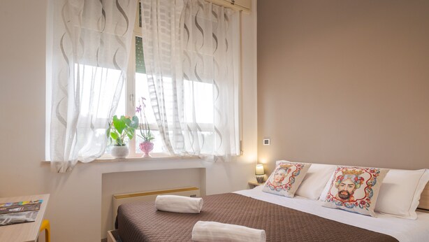 A bedroom with a bed, a window with sheer curtains, a bedside table with a lamp, and a potted plant. - Lady Hill (Taormina)