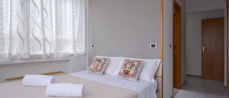 A hotel room with a bed, two pillows, a window with sheer curtains, a wooden door, and a tiled floor.