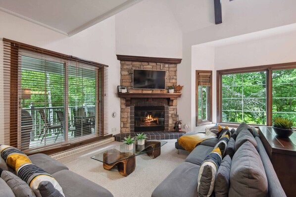 A living room with a stone fireplace, a sofa, a glass coffee table, and large windows with blinds. - Tremblant Prestige - Manoir 118-10 (Mont-Tremblant)