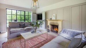 A living room with a fireplace, a sofa, a coffee table, and a large window. - Large family friendly holiday home in the Cotswolds - Willows House (Lower Swell)