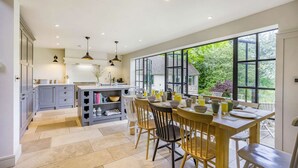 A modern kitchen with grey cabinets, a central island, and a dining area with wooden chairs and a table. - Large family friendly holiday home in the Cotswolds - Willows House (Lower Swell)