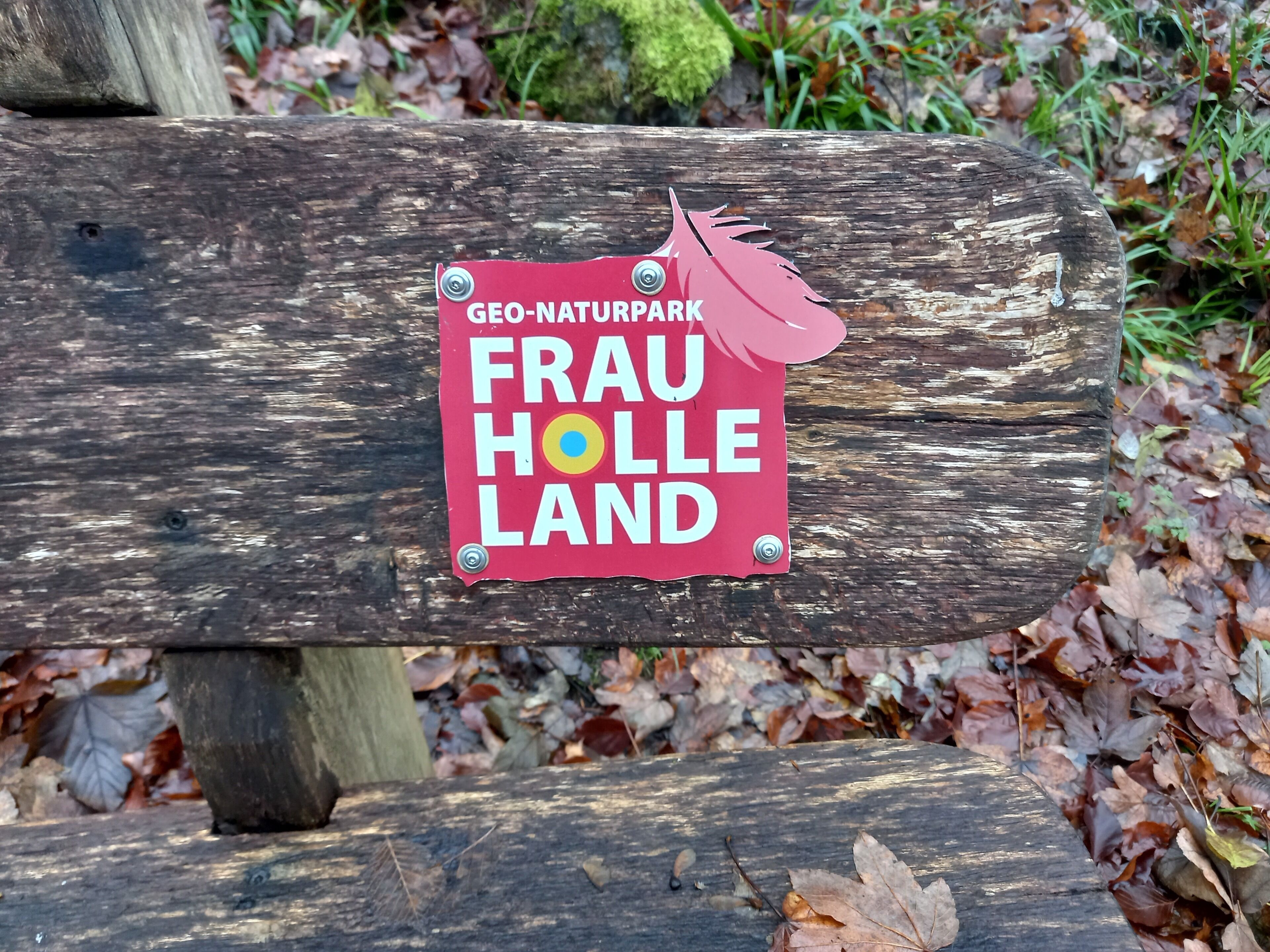 Un letrero de madera con etiqueta roja que dice 'Frau Holle Land' y una pequeña decoración de hoja rosa.